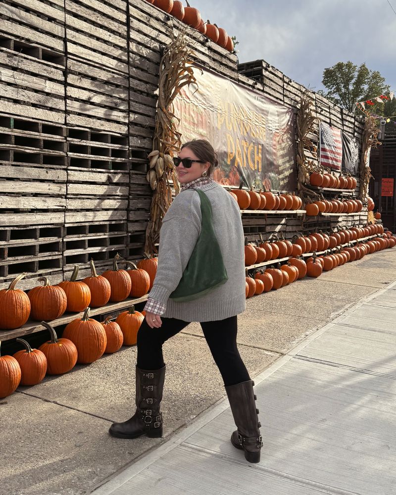 Maxey walking along a sidewalk past rows of orange pumpkins and a wall of stacked wooden pallets, looking back over her shoulder and smiling. She wears sunglasses, an oversized gray sweater over a plaid shirt, black leggings and tall brown buckled boots, with a green shoulder bag.